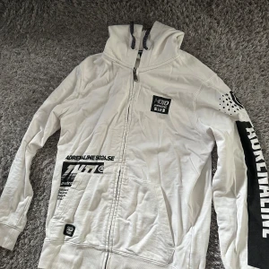 Vit hoodie från Petrol Industries - Vit hoodie från Petrol Industries med svart tryck och coola detaljer på ärmen. Tröjan har huva med snörning, dragkedja framtill och stora fickor. Perfekt för dig som gillar streetwear och vill sticka ut med en unik look.