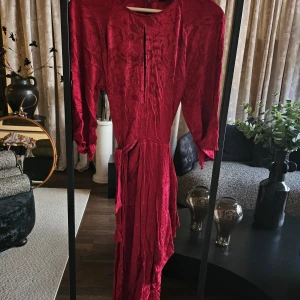 Vinröd långklänning med knytband - Säljer en vinröd långklänning med vackert strukturmönster och knytband i midjan. Klänningen har långa ärmar och en snygg slits framtill. Materialet har en lätt glans och känns lyxigt. Perfekt för dig som vill sticka ut med en elegant och unik look.