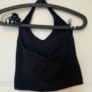 Svart ribbad halterneck croppad topp - Svart croppad halterneck topp med ribbad struktur och v-ringad hals. Snygg och enkel design som ger en cool vibe till din outfit. Perfekt att matcha med jeans eller kjol för en trendig look. Materialet känns mjukt och stretchigt.