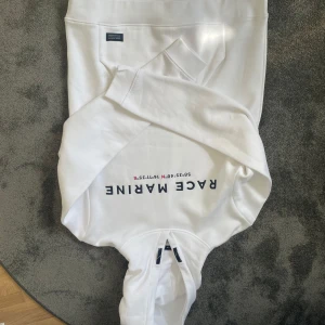 Vit hoodie från Tommy Hilfiger - Säljer en stilren vit hoodie från ’RACE MARINE' tryckt på framsidan. Hoodien har en klassisk design med en stor ficka framtill och en justerbar huva. Perfekt för en avslappnad look.