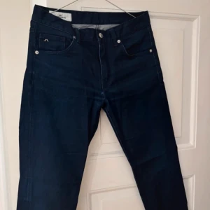 Mörkblå jeans från J.Lindeberg - Snygga mörkblå jeans från J.Lindeberg med klassisk femficksdesign. De har en rak passform och är tillverkade i ett slitstarkt denimtyg. Perfekta för en stilren look. Perfekt för att lyfta din outfit!