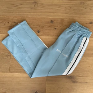 Ljusblå träningsbyxor från Palm Angels - Snygga ljusblå träningsbyxor från Palm Angels med elastisk midja och vita sidoremsor med svarta detaljer. Perfekta för en sportig look.