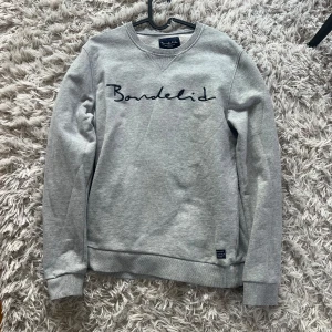 Grå sweatshirt från Bondelid - Säljer en stilren grå sweatshirt från Bondelid med broderad logga på bröstet. Tröjan har långa ärmar och ribbade muddar vid ärmslut och nederkant. Perfekt för en avslappnad look.