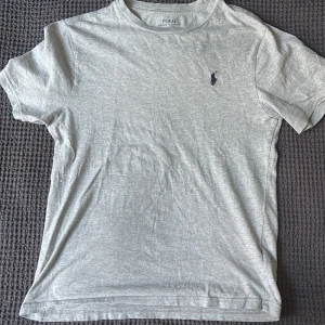 Grå t-shirt från Ralph Lauren - Säljer en stilren grå t-shirt från Ralph Lauren med korta ärmar.  Perfekt för en snygg outfit och passar i alla årstider. Tillverkad i mjukt bomullsmaterial för en skön och slät yta.