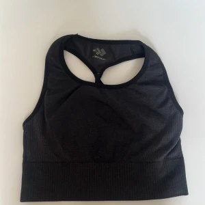 Svart sport-bh  - Säljer en svart sport-bh från Domyos med korsade band i ryggen. Perfekt för träning med sin bekväma passform och stretchiga material. Den har en ribbad kant nedtill för extra stöd.