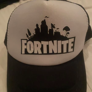 Svart och vit Fortnite keps - Cool svart och vit keps med Fortnite-tryck framtill. Perfekt för gaming-fans! Kepsen har ett nätmaterial baktill för extra ventilation och en justerbar rem för bästa passform.