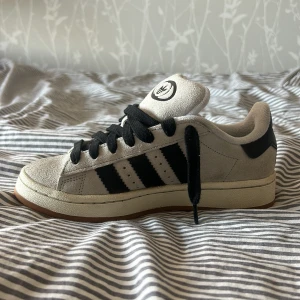Grå och svarta Adidas Campus sneakers - Snygga Adidas Campus sneakers i grå mocka med svarta detaljer och klassiska tre ränder. Skorna har en bekväm passform och svart snörning. Perfekta för en stilren look. Använda 3 gånger då storleken var för liten, har ett par vita snören också som såklart hänger med!💗 priset går såklart att diskutera.