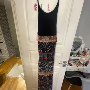 Mönstrad långklänning med spetsdetaljer - Säljer en bohemisk långklänning med mönster i flera färger. Klänningen har en svart överdel med spets och smala axelband som korsas i ryggen. Kjoldelen har ett färgglatt mönster med inslag av blommor och geometriska former.