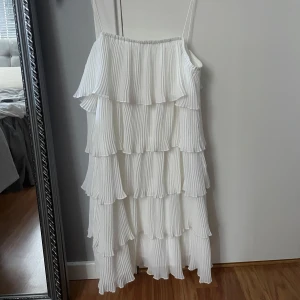 Vit volang klänning - Säljer en söt vit klänning med tunna axelband och flera lager av plisserade volanger. Perfekt för sommarens alla tillfällen. Klänningen har en luftig och romantisk känsla.