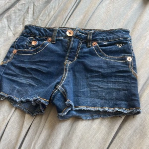 Blå jeansshorts från Justice - Snygga blå jeansshorts från Justice med fransiga kanter och dekorativa sömmar. Shortsen har flera fickor och knappar i kopparfärg. Perfekta för en avslappnad stil.