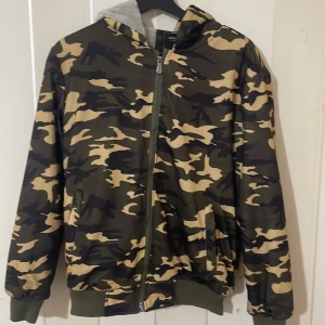 Camouflage bomberjacka med huva - Snygg bomberjacka i camouflage med dragkedja och huva. Jackan har ribbade muddar och är perfekt för en cool och avslappnad stil.