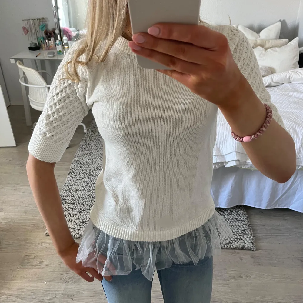 Säljer en stilren vit ribbad topp med trekvartsärm och spetsdetaljer på ärmarna. Toppen har en unik design med en volangkant i tyll vid nederkanten. Perfekt för att ge en romantisk touch till din outfit.. Neuletakit & Villapaidat.