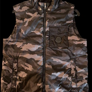 Camouflageväst från Moncler - Snygg camouflageväst från Moncler med dragkedja och praktiska fickor. Perfekt för en cool och avslappnad stil. Västen har en klassisk design med Moncler-logga på bröstfickan.