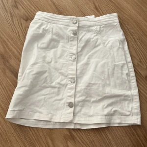 Vit kjol med knappar från H&M - Säljer en stilren vit kjol från H&M med knappar framtill. Kjolen har en klassisk design med fickor och är perfekt för en somrig look. Passar bra till både vardag och speciella tillfällen.