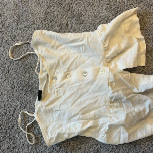 Vit ärmlös blus med knappar - Säljer en stilren vit blus med knappar framtill. Den är ärmlös och har en luftig passform, perfekt för varma dagar. Blusen har smala axelband och en lätt rynkad detalj vid midjan.