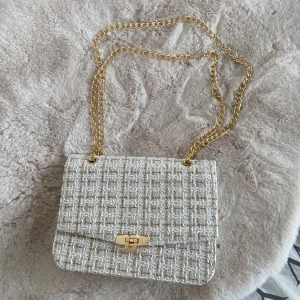 Vit och beige axelväska med kedja - Snygg axelväska i vit och beige med ett rutigt mönster. Väskan har en elegant guldkedja och ett stilrent spänne framtill. Perfekt för att ge din outfit en chic touch.