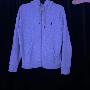 Grå Ralph Lauren zip - Säljer en stilren grå hoodie från Ralph Lauren med dragkedja och justerbar huva. Den har långa ärmar och en liten broderad logga på bröstet. Perfekt för en avslappnad look. Storleken är M/175/96A men passar mig som är 190cm.