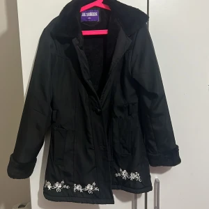 Svart kappa med broderi - Snygg svart kappa från Al Yaseen med vackra vita broderier längst ner. Jackan har en mjuk pälsliknande insida och långa ärmar. Perfekt för kyligare dagar.