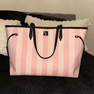  Victoria's Secret Axel väska - Snygg axelväska från Victoria's Secret i rosa och vit randigt mönster. Väskan har svarta handtag och detaljer, samt en elegant V-logga framtill. Perfekt för att bära med sig allt man behöver med stil. Dessutom svart tyg inuti hela där det står Victorias secret, jätte fint verkligen! (KNAPPT ANVÄND)💖.                          OPS! Går ej att få tag på i Sverige (går inte heller att beställa) har man tur så finns den utomlands. Denna är köpt i Barcelona🥰