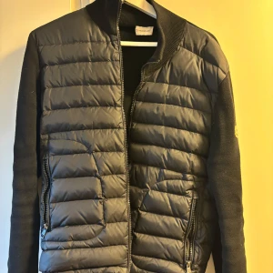 Svart Moncler cardigan  - Snygg svart cardigan .Jackan har en stilren design med Moncler-logga på ärmen. Perfekt för kyligare dagar. Säljer den för att den är för liten för mig , den är sparsamt använd.