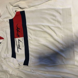 Vit tröja från Tommy Hilfiger - Säljer en stilren vit tröja från Tommy Hilfiger med ett färgglatt tryck i rött, vitt och blått på bröstet. Tröjan har långa ärmar och en klassisk passform. Perfekt för en avslappnad stil. Den passar som en xs/s