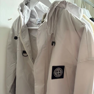Vit tunn jacka från Stone Island  - Säljer en stilren vit jacka från Stone Island med huva och dragkedja. Jackan har en ikonisk logga på bröstet och justerbara snören vid huvan. Perfekt för en minimalistisk look.