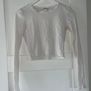 Vit ribbad croppad topp från Monki  - Säljer en stilren vit ribbad croppad topp från Monki. Toppen har långa ärmar och en rund halsringning, perfekt för lager-på-lager eller som den är. Passar till många olika outfits!