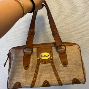 Beige och brun handväska från XOXO - Snygg handväska från XOXO i beige med bruna detaljer. Väskan har ett mönster med XOXO-loggan och är försedd med dragkedja. Den har två handtag och dekorativa nitar framtill. Perfekt för att bära dina dagliga nödvändigheter med stil.