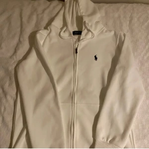 Vit hoodie från Ralph Lauren - Säljer en stilren vit hoodie från Ralph Lauren med dragkedja och det klassiska logotypbroderiet på bröstet. Perfekt för en avslappnad look. Den har långa ärmar och en bekväm huva.