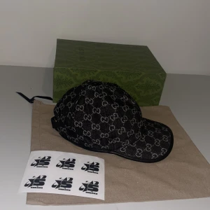Svart keps från Gucci - Snygg svart keps från Gucci med det ikoniska GG-mönstret i grått. Kepsen har en justerbar rem baktill för perfekt passform. Perfekt för att ge din outfit en lyxig touch. Finns i storlek S M L