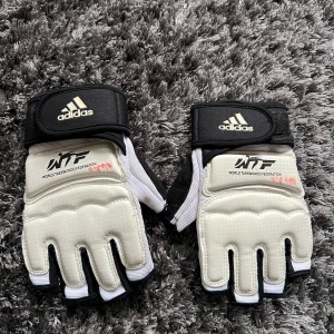 Adidas Taekwondo handskar - Säljer ett par Adidas Taekwondo handskar i vitt och svart. De har en öppen design för fingrarna och kardborreband för justering. Perfekta för träning och tävling inom kampsport.