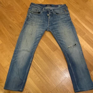 Blå jeans från Replay - Snygga blå jeans från Replay med en sliten look och en reva på ena benet. Modellen är Newbill och de har en klassisk femficksdesign med knappgylf.