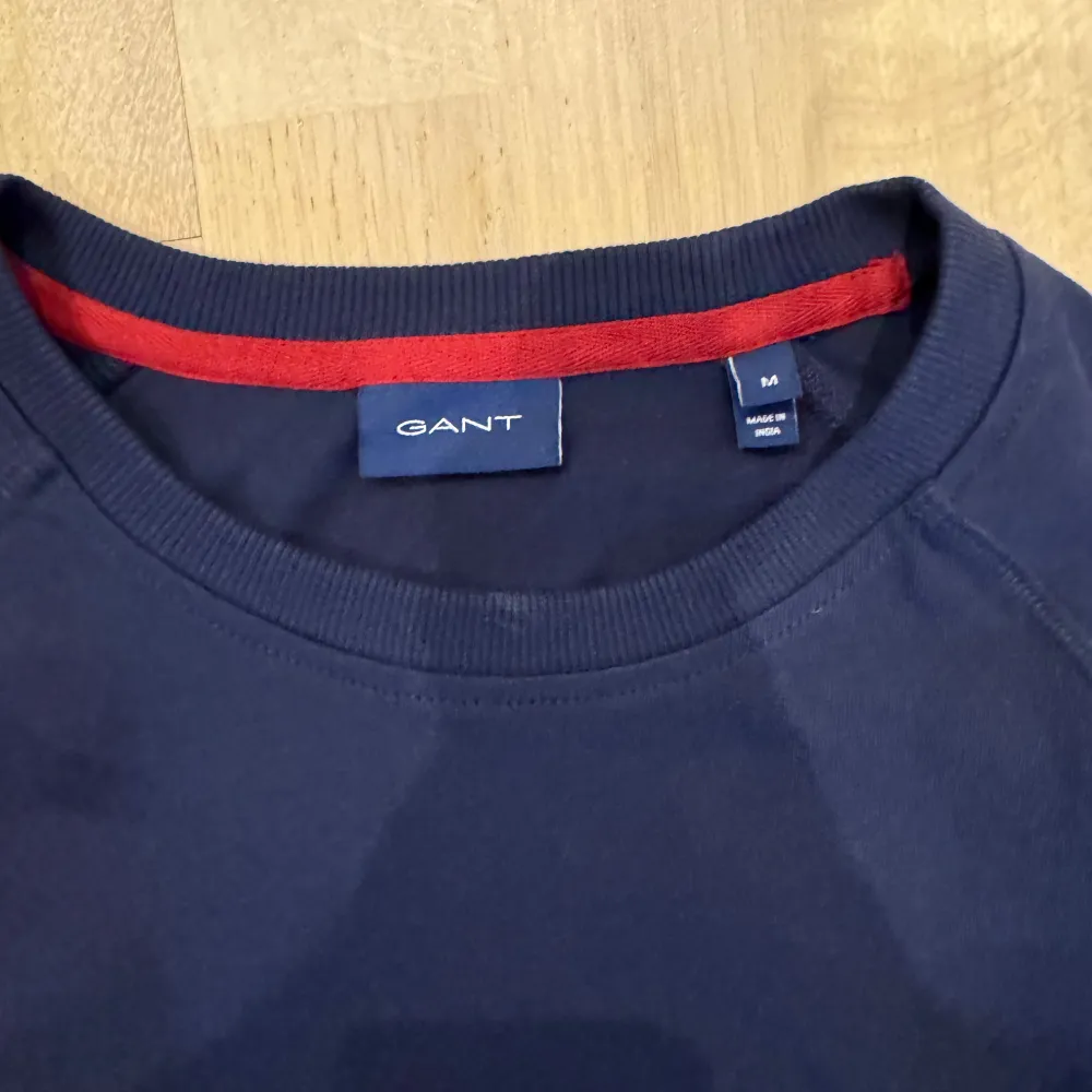 Säljer en mörkblå sweatshirt från GANT med vit panel och röd text 'GANT 1949' på bröstet. Tröjan har långa ärmar och ribbade muddar. Perfekt för en avslappnad stil.. Neuletakit & Villapaidat.