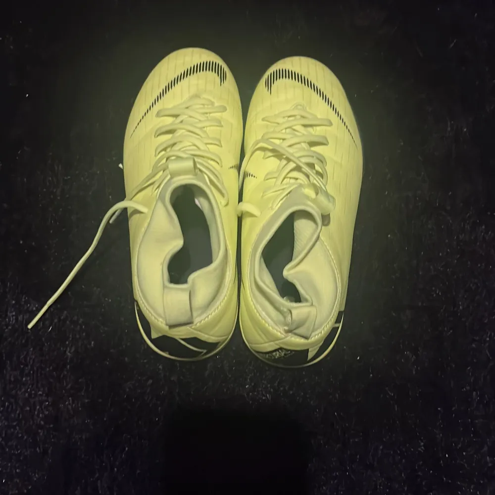 Snygga gula fotbollsskor från Nike med snörning och hög ankel. Skorna har en modern design med svarta detaljer och en slitstark sula för bra grepp på planen. Perfekta för den som vill sticka ut på fotbollsplanen.. Kengät.