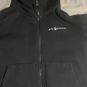 Svart jacka från Sail Racing - Snygg svart jacka från Sail Racing med dragkedja och huva. Perfekt för en stilren look. Jackan har en enkel design med logotyp på bröstet och huvan.