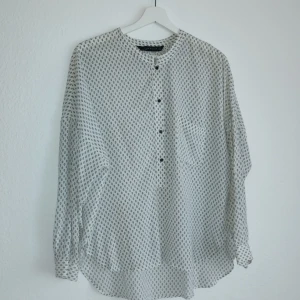 Vit och svart mönstrad blus från Zara Woman - Säljer en stilren vit blus från Zara Woman med svart geometriskt mönster. Blusen har en henley-krage och knappar framtill. Perfekt för en chic och avslappnad look.