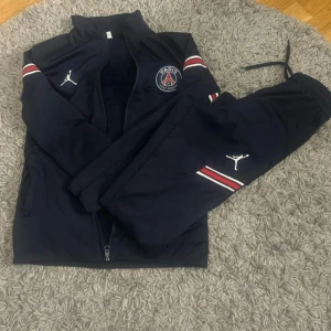 Mörkblå träningsset från Jordan x PSG - Snyggt mörkblått träningsset från Jordan i samarbete med PSG. Setet har vita och röda detaljer samt PSG-logga på både jacka och byxor. Jackan har dragkedja och byxorna har resår i midjan för en bekväm passform.