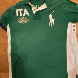 Grön pikétröja från Ralph Lauren - Säljer en grön pikétröja från Ralph Lauren med vita detaljer och broderad logga. Tröjan har korta ärmar och knappar vid kragen. Texten 'ITA' och 'ITALY' samt siffran '2' finns på framsidan och baksidan.