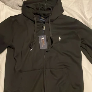 Svart hoodie från Polo Ralph Lauren - Säljer en stilren svart hoodie från Polo Ralph Lauren med dragkedja och den klassiska broderade loggan på bröstet. Perfekt för en avslappnad look. Hoodien har snörning i huvan och är tillverkad i ett mjukt material.