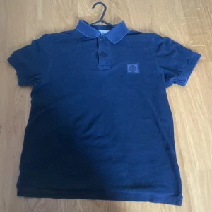 Mörkblå pikétröja från Stone Island - Säljer en snygg mörkblå pikétröja från Stone Island. Tröjan har en klassisk krage med knappar och en diskret logotyp på bröstet. Perfekt för en stilren look.