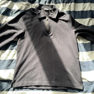 Svart långärmad pikétröja från Dobber - Snygg svart långärmad pikétröja från Dobber med dragkedja vid halsen. Perfekt för en stilren och avslappnad look. Tröjan har en klassisk krage och är tillverkad i ett bekvämt material.