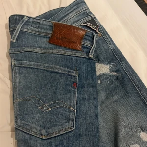 Blå jeans från Replay - Snygga blå jeans från Replay med en klassisk femficksdesign och en sliten look. De har en läderpatch med Replay-logga på baksidan och en liten röd detalj vid fickan. Perfekta för en avslappnad stil.
