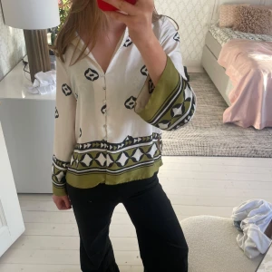 Vit och grön mönstrad blus från Zara  - Snygg vit blus med gröna och svarta mönsterdetaljer. Blusen har långa ärmar och knappar framtill. Perfekt för en stilren look.