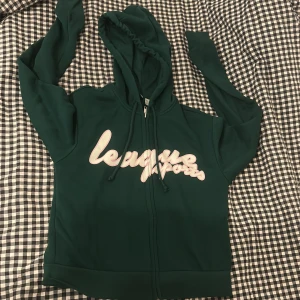 Grön hoodie från H&M Divided - Säljer en grön hoodie från H&M Divided med texten 'League Sports' i rosa på framsidan. Den har en dragkedja och en bekväm huva med snören. Perfekt för en avslappnad stil.