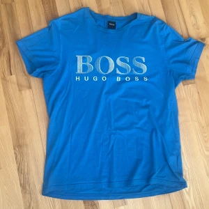 Blå t-shirt från Hugo Boss - Säljer en snygg blå t-shirt från Hugo Boss med stort BOSS-tryck på framsidan. T-shirten är kortärmad och perfekt för en avslappnad stil. Passar bra till jeans eller shorts för en cool look. T-shirten är helt ny och bara använd 2 gånger