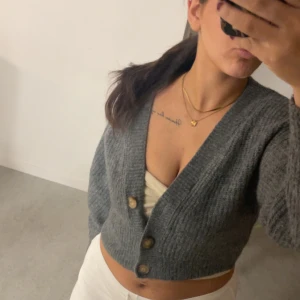 Grå stickad kofta - Säljer en mysig grå stickad kofta med stora knappar framtill. Koftan är croppad och har långa ärmar, perfekt för en avslappnad stil. Passar bra till både jeans och kjol. Hör av dig vid intresse!