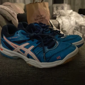 Blå träningsskor från Asics - Säljer ett par blå träningsskor från Asics med vita och orange detaljer. Skorna har snörning och en slitstark sula i gummi. Perfekta för inomhusträning eller sportaktiviteter.