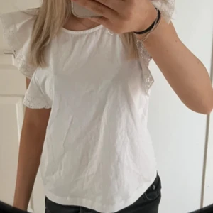 Vit blus med volangärmar - Säljer en söt vit blus med volangärmar och spetsdetaljer. Perfekt för en somrig look. Blusen har en rund halsringning och en luftig passform.