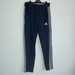 Blå träningsbyxor från Adidas - Snygga blå träningsbyxor från Adidas med de klassiska tre vita ränderna längs sidorna. Byxorna har en elastisk midja med dragsko för bästa passform och komfort. Perfekta för träning eller en avslappnad dag.