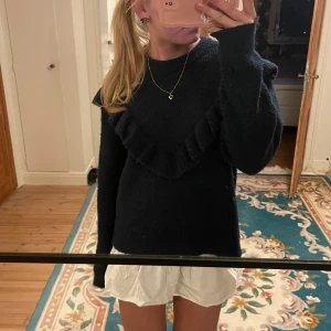 Mörkblå stickad tröja med volang - Säljer en snygg mörkblå stickad tröja med volangdetaljer över bröstet. Tröjan har långa ärmar och en bekväm passform, perfekt för kyligare dagar. Passar bra till både jeans och kjol. Perfekt för dig som vill ha något stilrent men ändå med en twist.